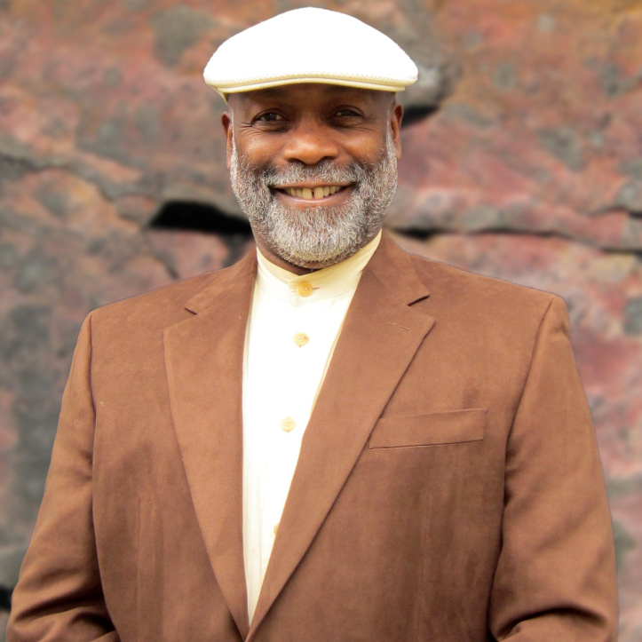 Lawrence Diggs Photograph Image of artist Lawrence Diggs. Diggs is shown from the waist up, wearing a brown suit jacket, off-shite collared shirt, and white cap. He is smiling through a full grey beard.