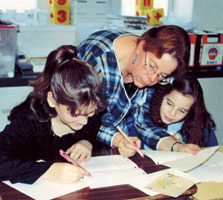 Photo of artist Artist helps two children with their artwork at a table.