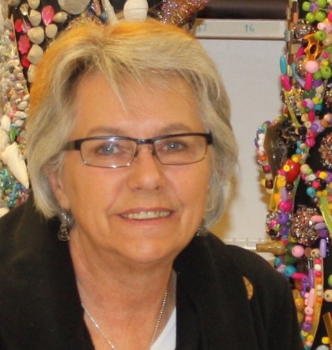 Photo of artist A person wearing a black jacket and a white shirt, stands in front of a colorful background filled with beads and jewelry items.