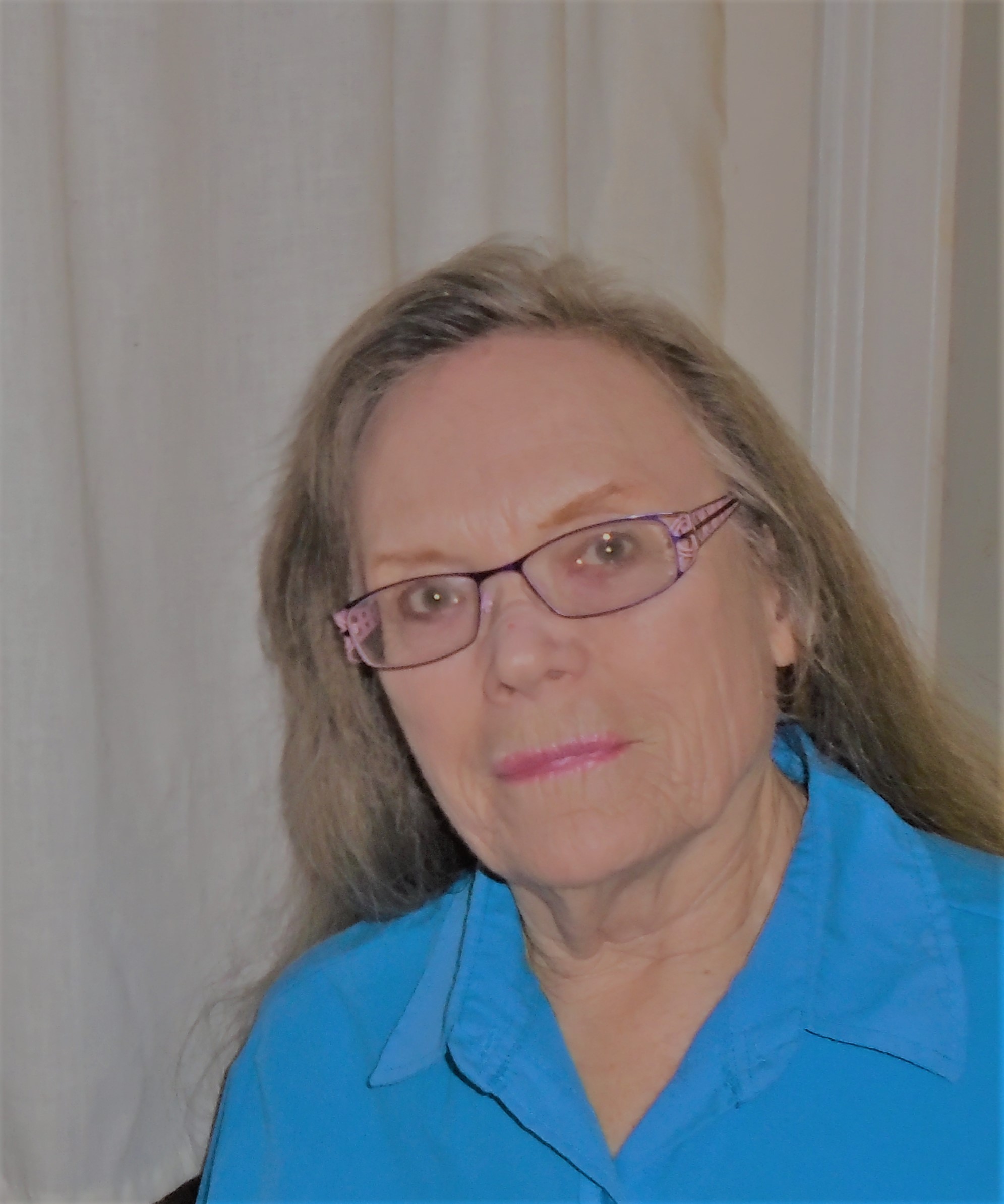 Photo of artist Artist wearing a blue collared shirt and pink-framed glasses, stands in front of a light-colored curtain.
