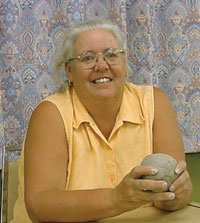 Photo of artist Artist sits at a table, holding a lump of clay with both hands, with a patterned curtain in the background.
