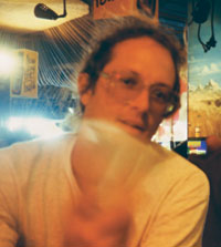 Photo of artist Artist wearing a light-colored shirt, with various objects in the background including a hanging light fixture and a sign with text that is not fully visible.