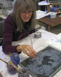 Photo of artist Artist manipulating paint or ink on the surface of water or a similar liquid medium in a tray, surrounded by jars of paint or ink and brushes.