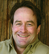 Photo of artist Artist wears a light-colored collared shirt, stands in front of a wooden surface with vertical lines.