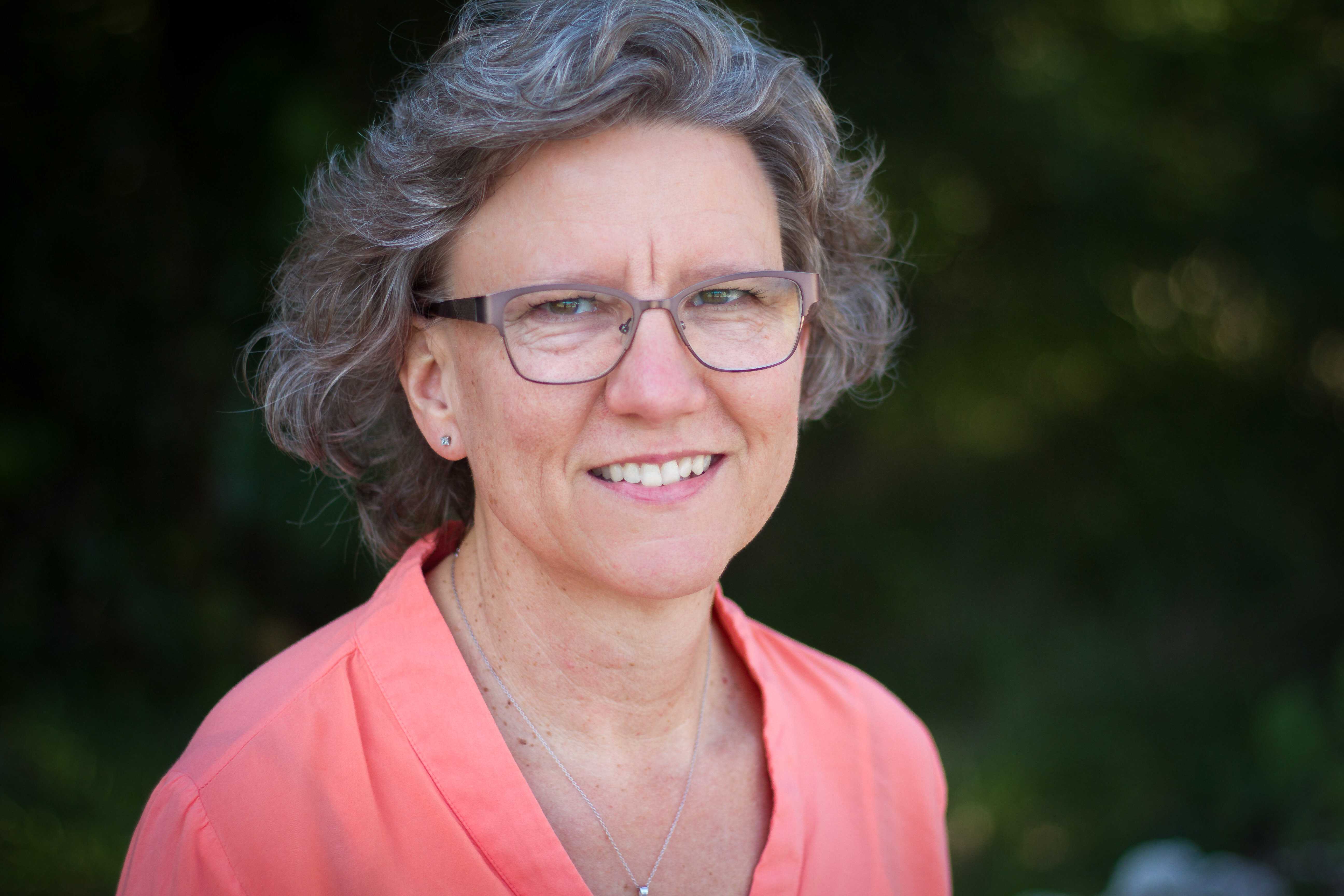 Photo of artist A person with short, curly gray hair wearing a coral-colored shirt and a necklace with a small pendant stands outdoors with blurred greenery in the background.