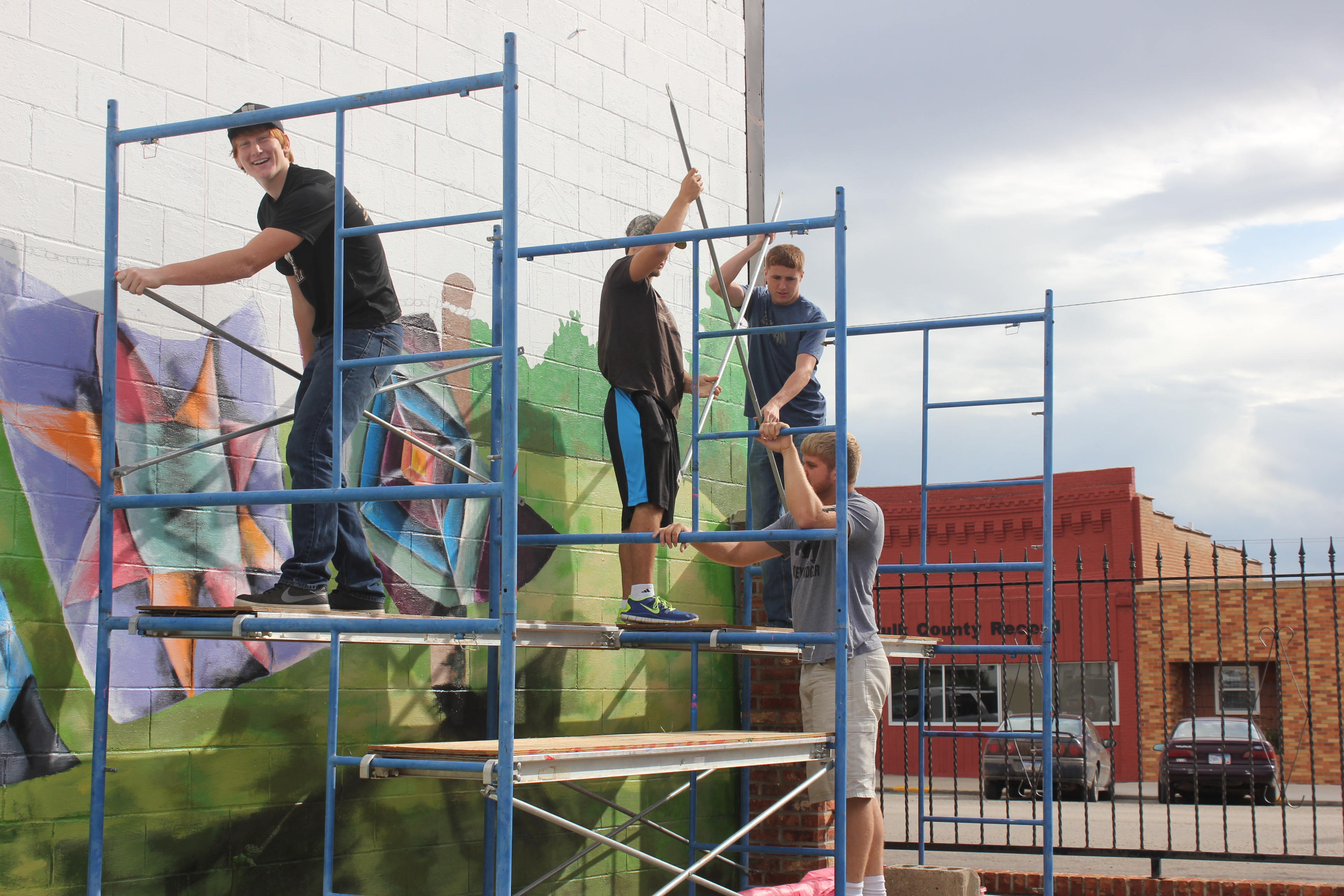 Youth Painting Mural on Brick Exterior Wall Image of young boys painting a brick exterior wall on a downtown main street. Four young men are standing on a scaffolding - one faces the camera while painting, two work with large rollers to get more paint, and one stands at the far end of the scaffold.